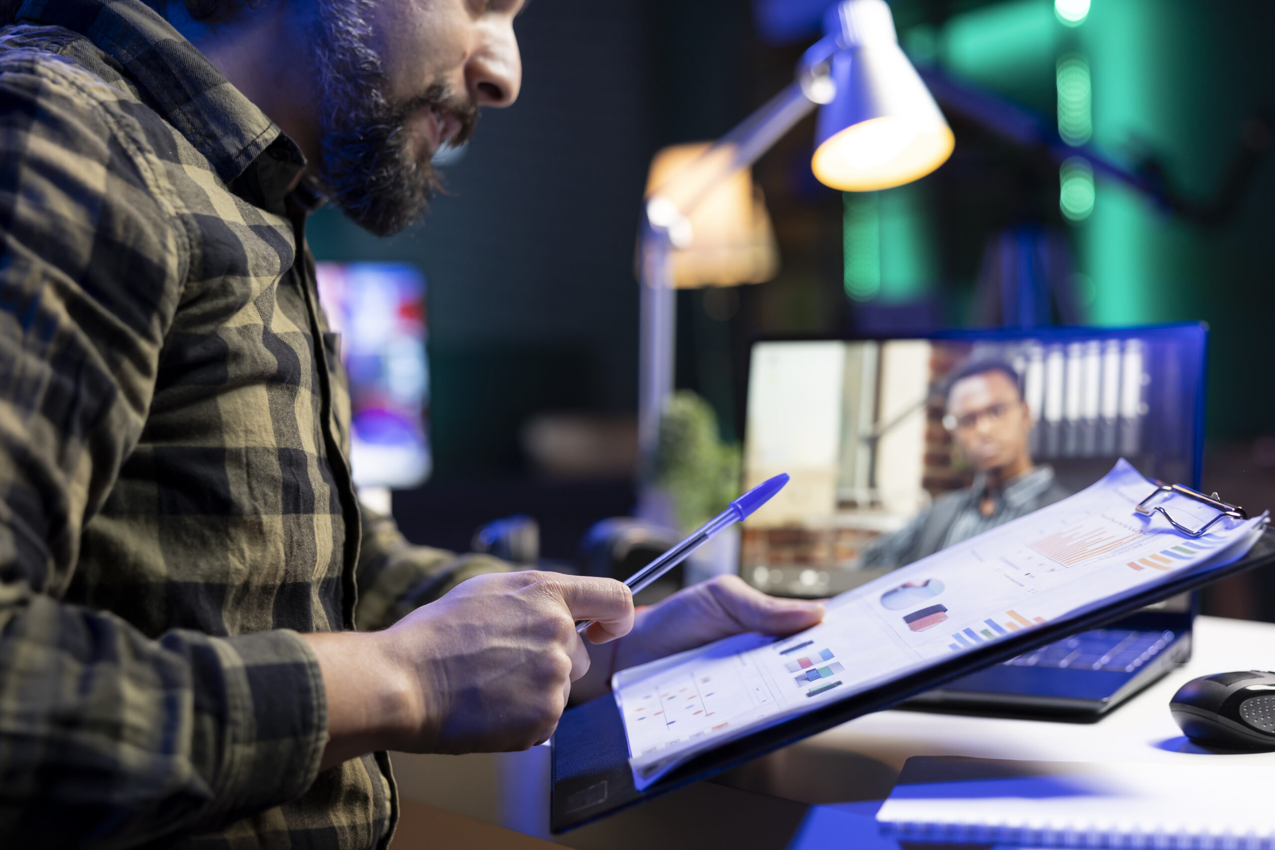 Bearded man with a pen and clipboard participating in virtual meeting, comparing data with african american person on laptop. Male individual having a video call while reviewing documents.